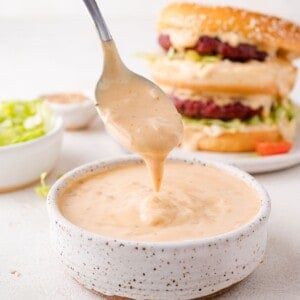 A spoon dripping creamy big mac sauce over a bowl. A hamburger with lettuce is in the background.