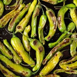A pile of blistered green shishito peppers with charred spots on a dark surface.