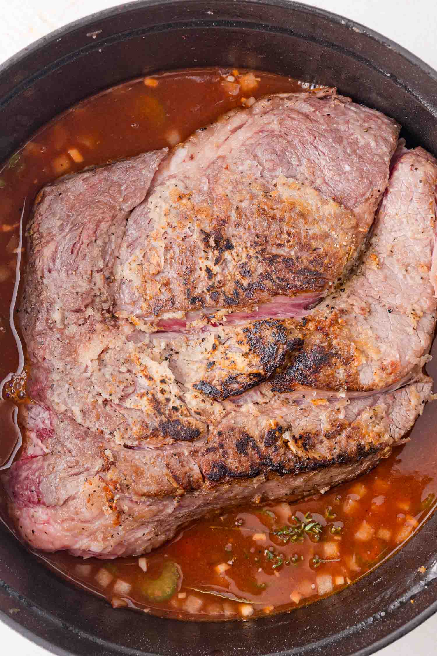 Browned beef roast in a black pot with sauce and diced onions.