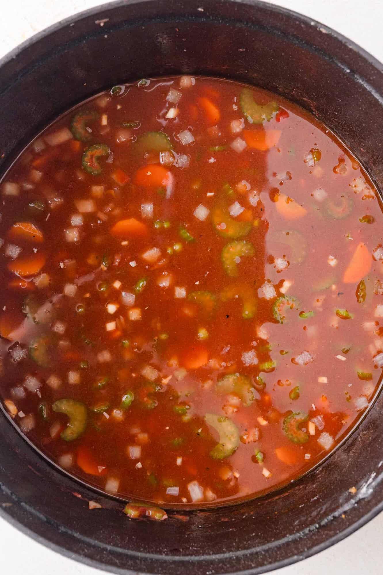 A pot containing visible pieces of carrots, celery, and onions in a reddish broth.