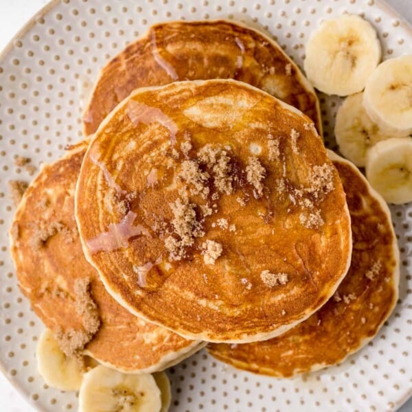 A plate of pancakes topped with brown sugar and syrup, surrounded by banana slices.