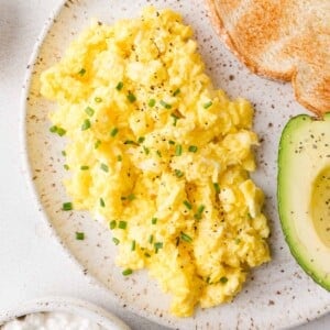 Cottage cheese scrambled eggs garnished with chives, served with sliced avocado and toasted bread on a speckled plate.