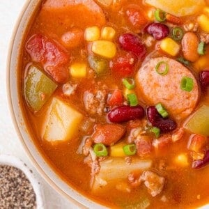 A bowl of hearty stew with sausage slices, beans, corn, potatoes, and diced vegetables garnished with chopped green onions.