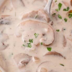 Close-up of creamy mushroom soup with sliced mushrooms and a garnish of chopped herbs, with a spoon partially visible.