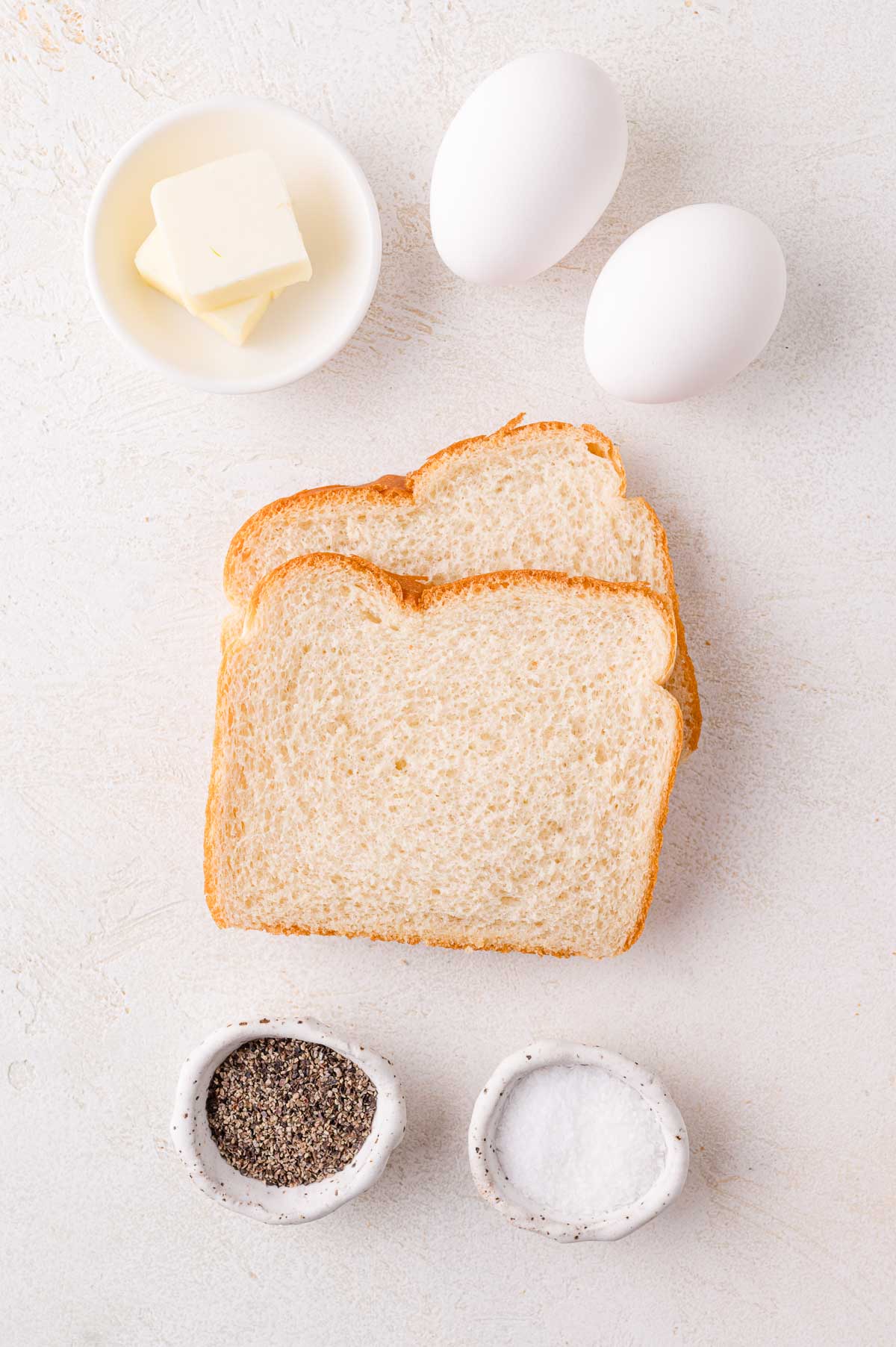 Two slices of bread, two eggs, a small dish of butter, a bowl of salt, and a bowl of pepper on a light surface.
