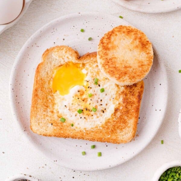 A piece of toast with a cooked egg in the center, garnished with chopped chives, served on a white plate with a toasted bread circle on the side.