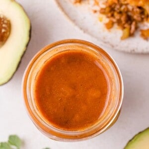 A jar filled with red enchilada sauce is centered on a table. Sliced avocado and a plate with food are partially visible nearby.