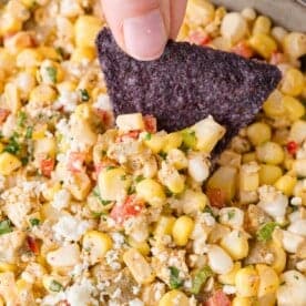A blue tortilla chip is dipped into a mixture of corn, diced red and green onions, and crumbled cheese.
