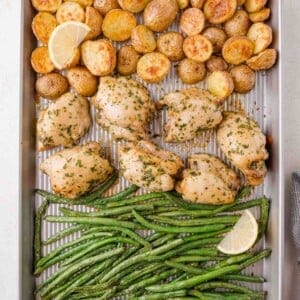 Sheet pan with roasted chicken, halved potatoes, and green beans, garnished with lemon wedges.