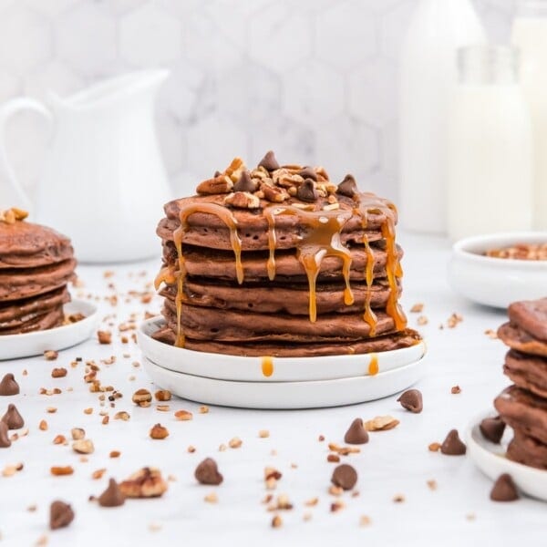 Stack of chocolate pancakes drizzled with caramel and topped with chopped nuts and chocolate chips, on a white plate. Nearby are more pancakes, a white jug, and a bottle.