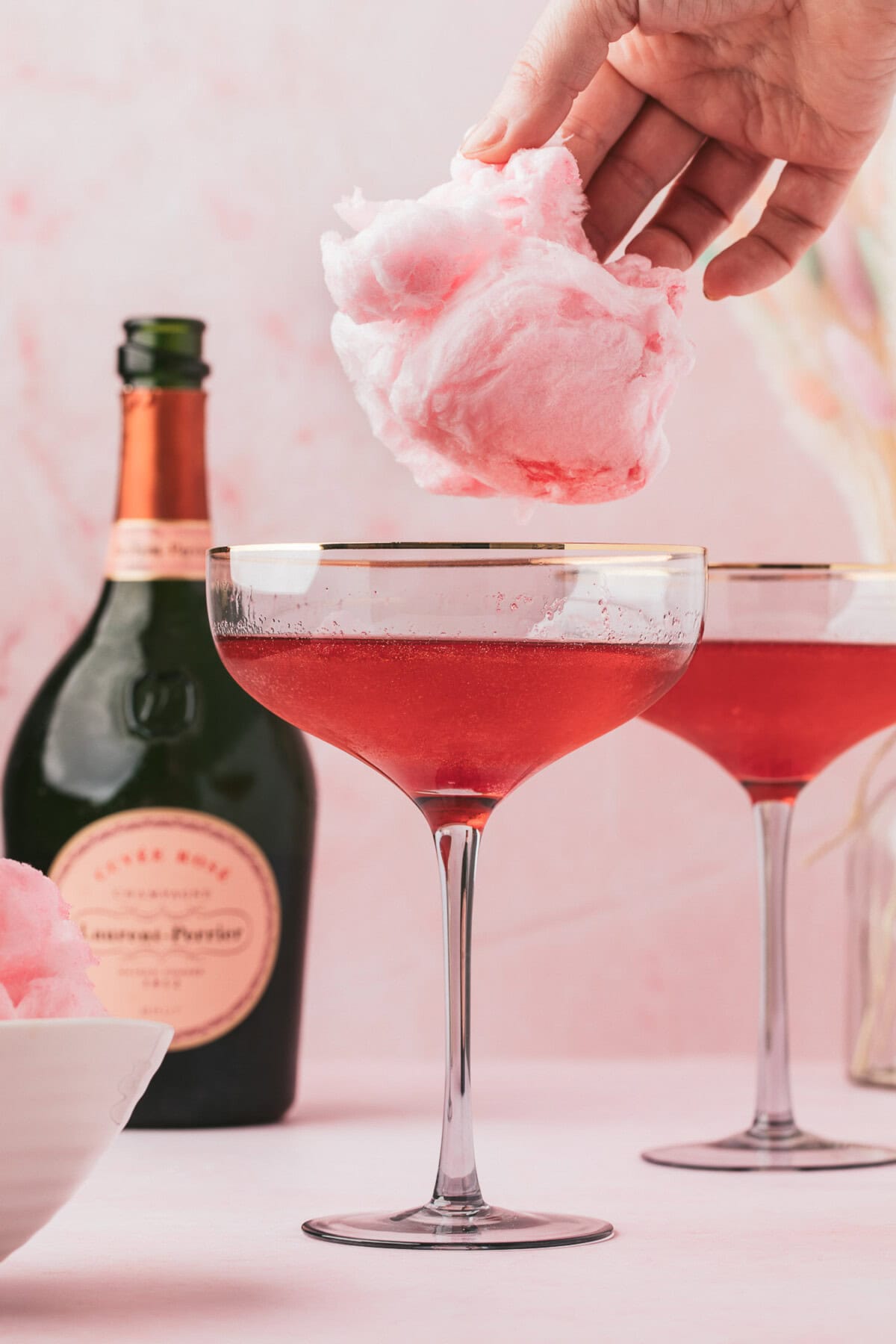 Cotton Candy Cocktail 5 A hand places pink cotton candy above a glass of red cocktail. A bottle of sparkling rosé and another cocktail glass are in the background.