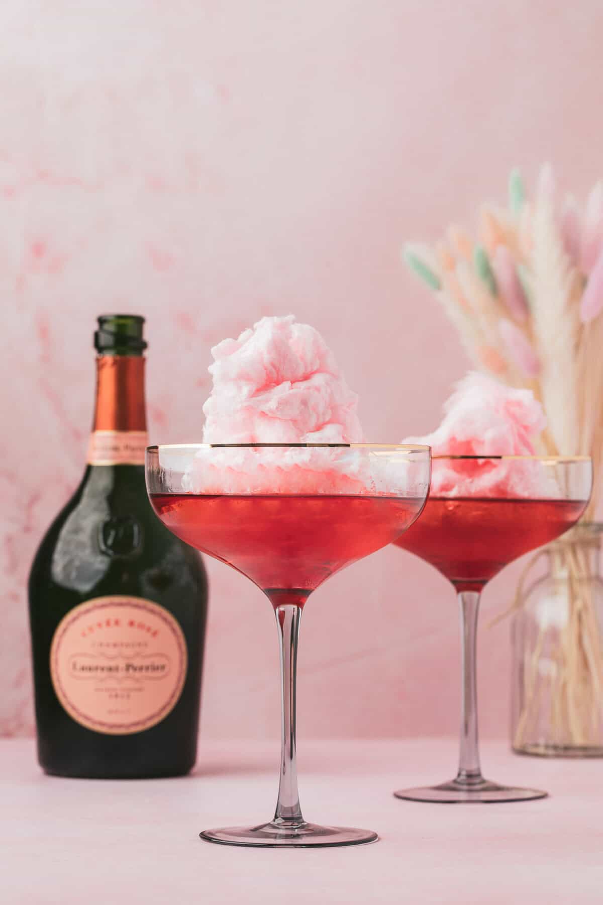 Cotton Candy Cocktail 6 Two cocktail glasses filled with pink drinks and topped with cotton candy, set on a pink surface.
