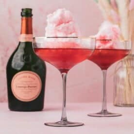 Two cocktail glasses filled with pink cotton candy and a pink liquid, placed next to a bottle of Laurent-Perrier champagne on a pink surface.