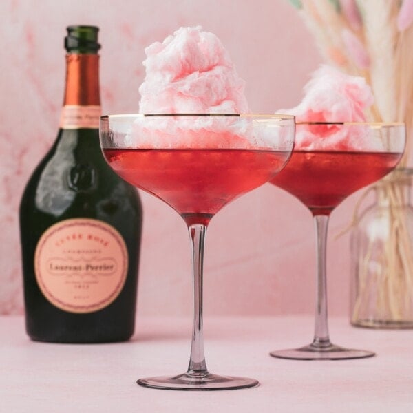 Two cocktail glasses filled with pink cotton candy and a pink liquid, placed next to a bottle of Laurent-Perrier champagne on a pink surface.