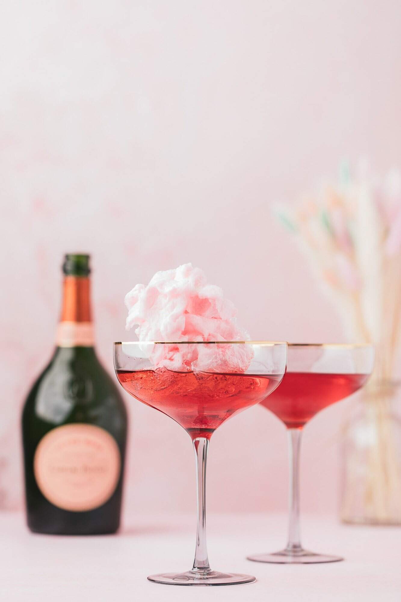 Cotton Candy Cocktail 2 Two glasses of pink cocktails with cotton candy garnish. A bottle of sparkling wine is in the background against a soft pink backdrop.