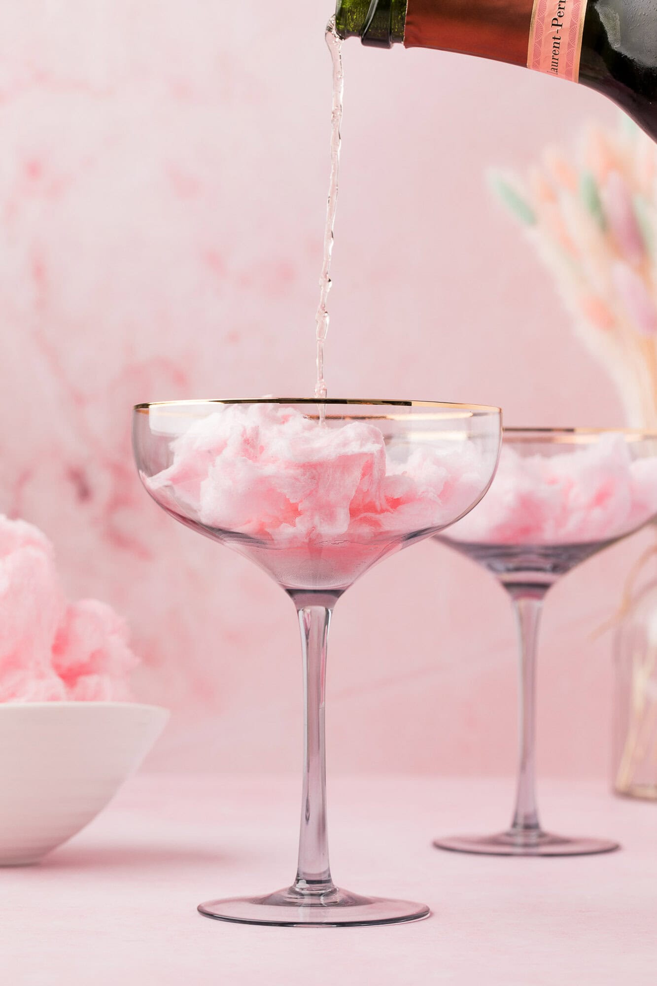Cotton Candy Cocktail 3 Champagne being poured into a glass filled with pink cotton candy, with another similar glass and a bowl of cotton candy in the background, set against a pink backdrop.
