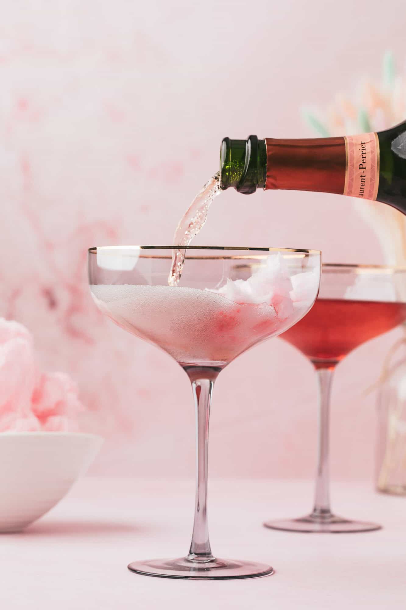 Cotton Candy Cocktail 4 Champagne being poured into a glass with cotton candy inside, with another similar drink and a bowl of cotton candy in the background.