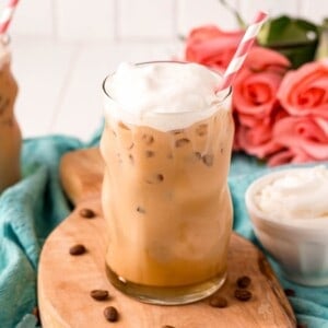 A glass of iced vanilla coffee with whipped cream and a striped straw on a wooden board, surrounded by coffee beans and pink roses in the background.