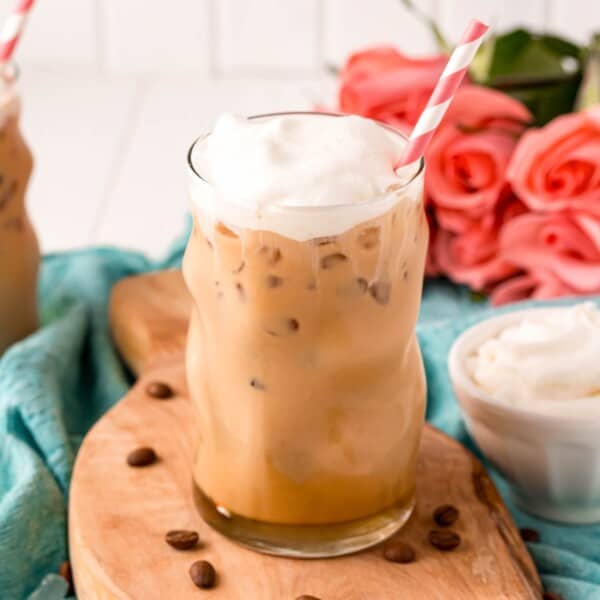 A glass of iced vanilla coffee with whipped cream and a striped straw on a wooden board, surrounded by coffee beans and pink roses in the background.