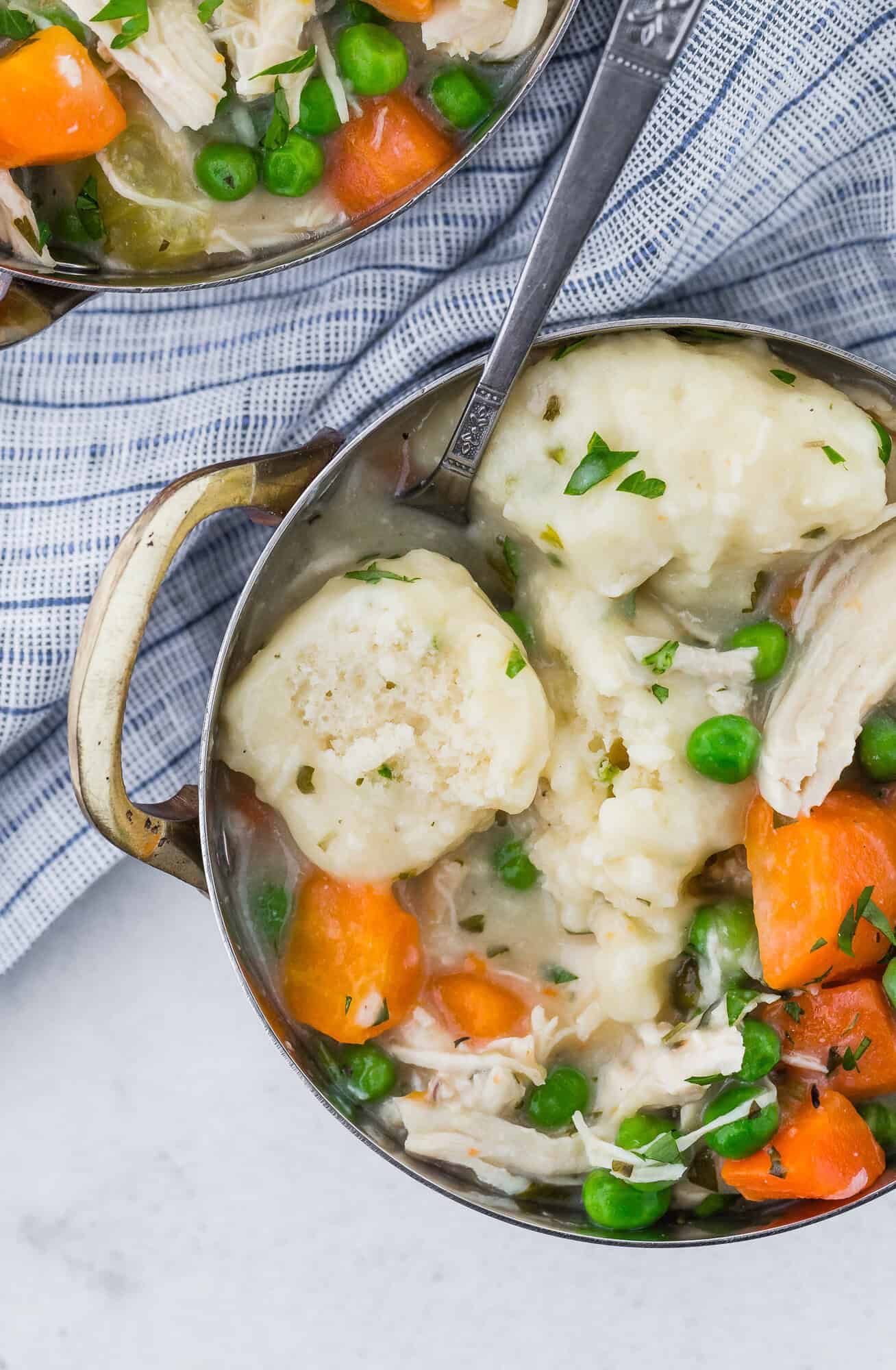 Bowl of chicken and dumplings with peas and carrots, garnished with herbs, on a striped cloth.