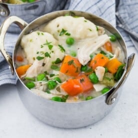 A metal bowl filled with chicken and dumplings, featuring carrots, peas, and parsley garnish on a gray tablecloth.