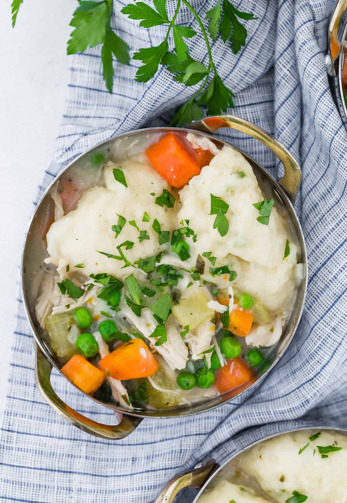 A bowl of Instant Pot chicken and dumplings with peas, carrots, and parsley garnish, set on a striped cloth.