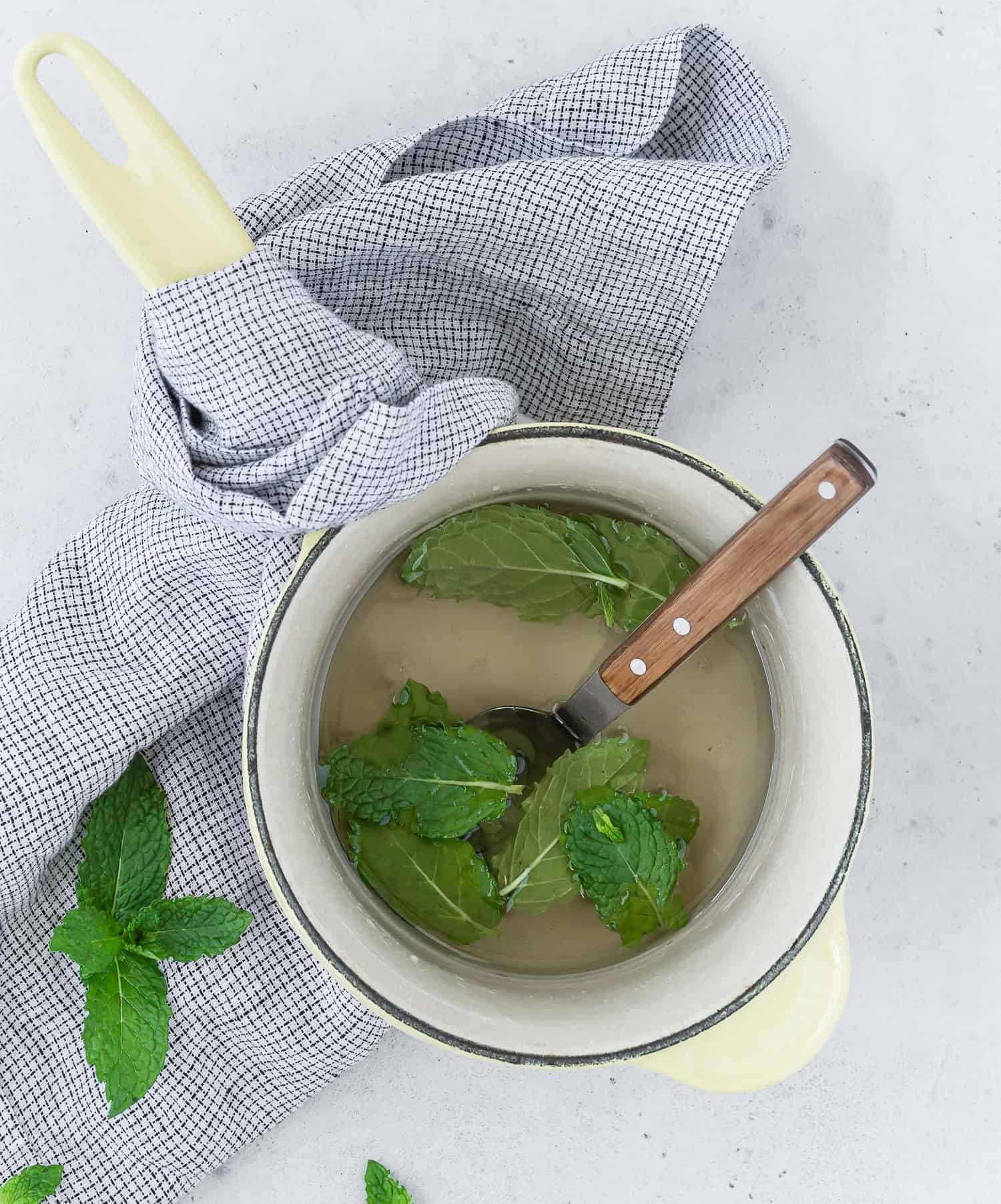 A saucepan with a wooden-handled spoon contains a liquid and mint leaves, resting on a gray checked cloth. Additional mint leaves and the cloth are on a light gray surface.