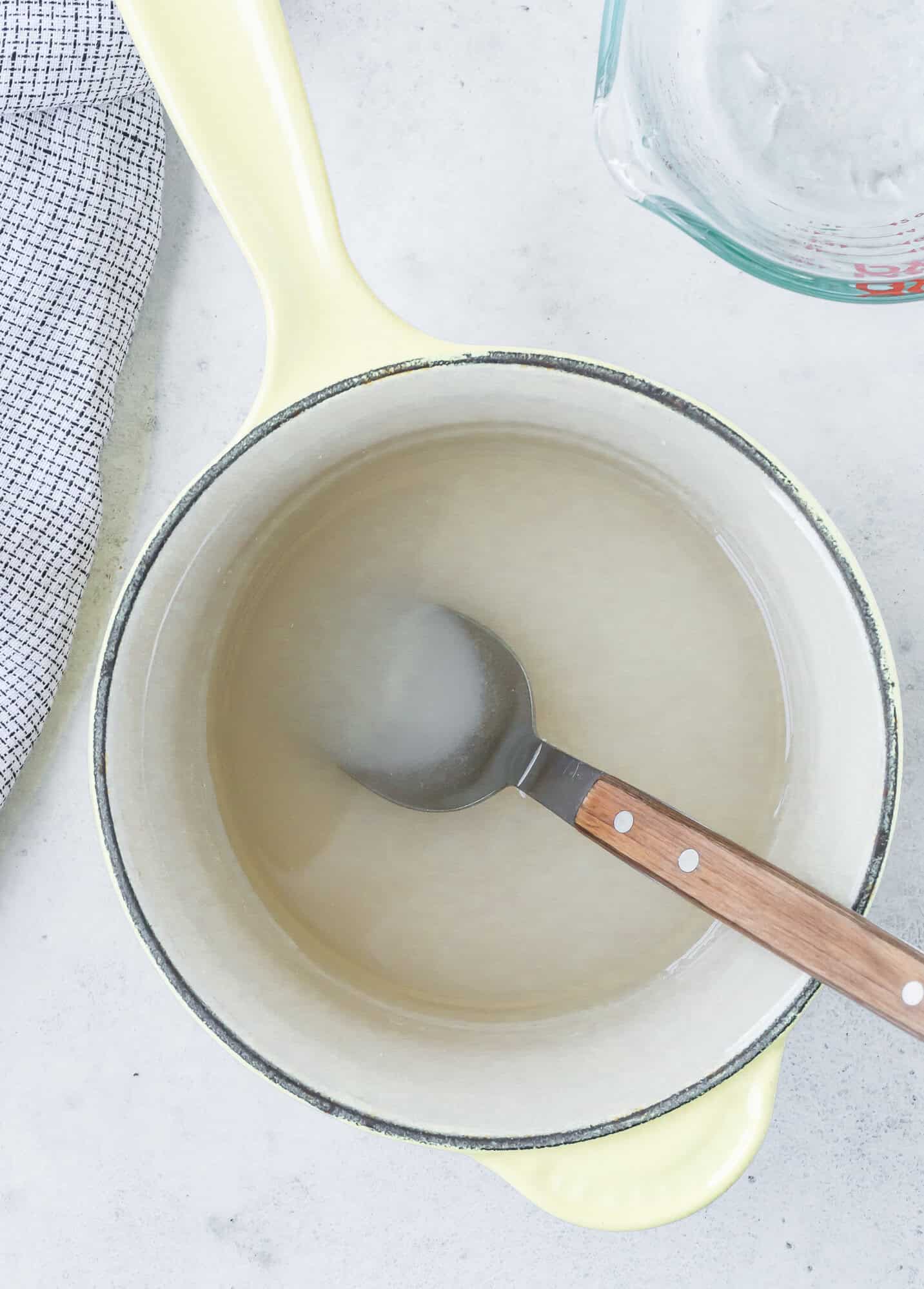A spoon rests in a pot containing a translucent liquid, with a measuring cup nearby and a checkered cloth to the side.