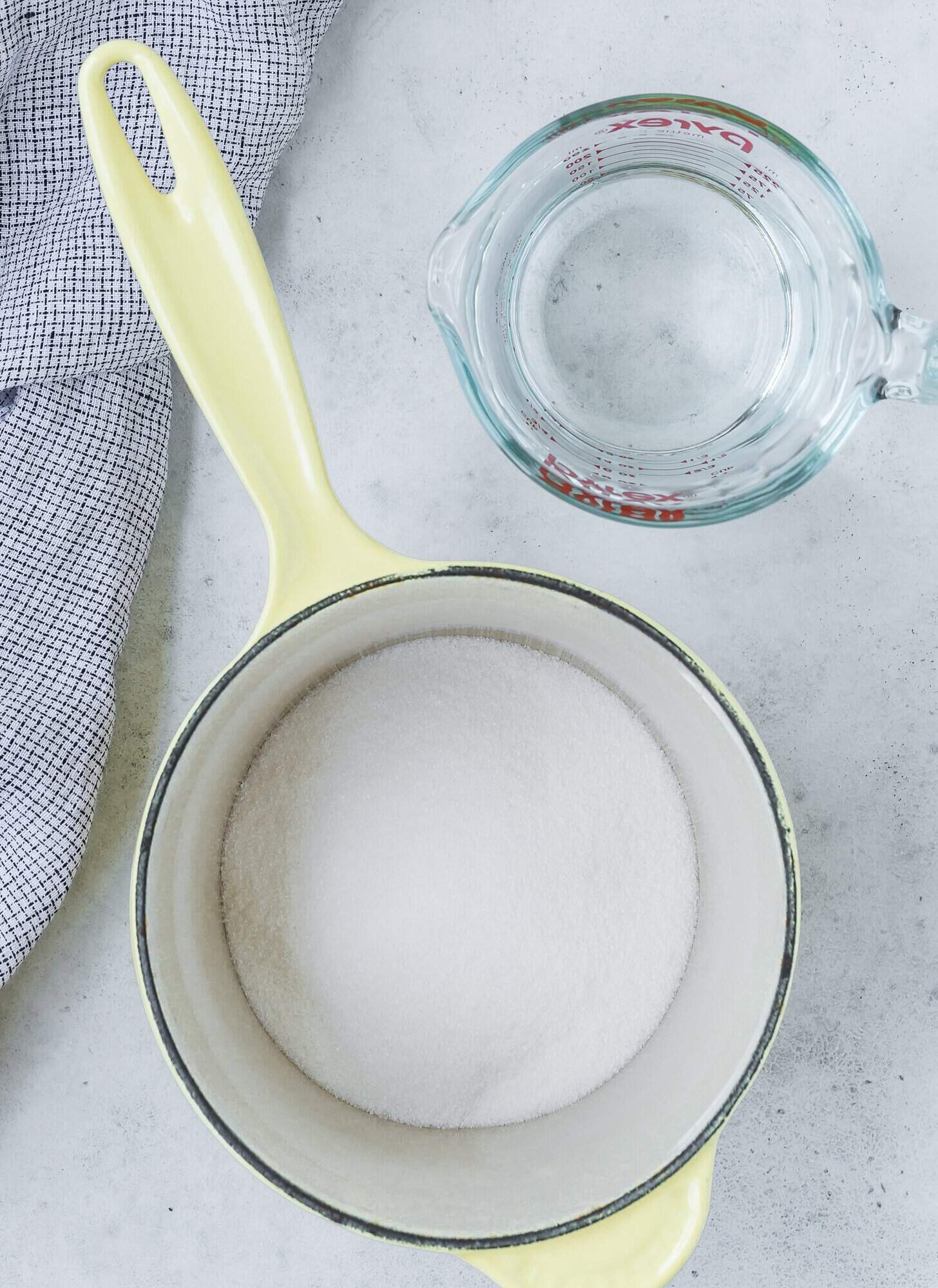 A pot with sugar inside next to a glass measuring cup of water on a countertop with a checkered cloth.