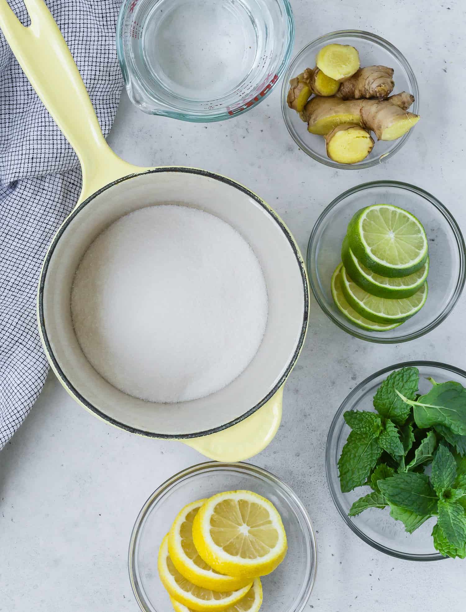 Ingredients for a infused simple syrup: lemon slices, lime slices, ginger pieces, mint leaves, a pot with sugar, and a measuring cup with water on a countertop.