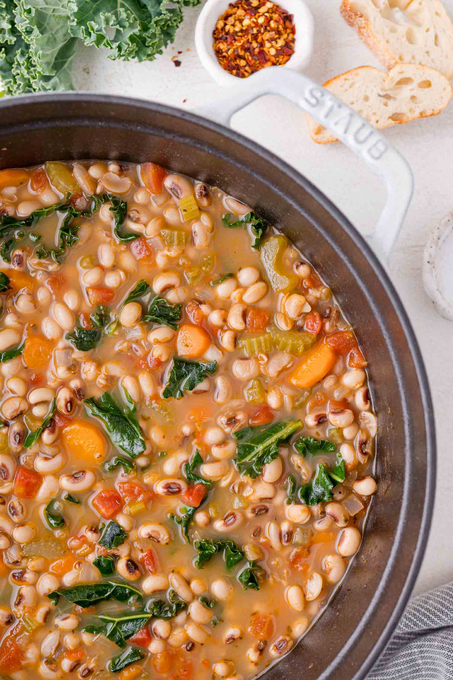 Black-Eyed Pea Soup 2 A pot of black-eyed pea soup with carrots, kale, and white beans, surrounded by slices of bread and a small bowl of red pepper flakes.