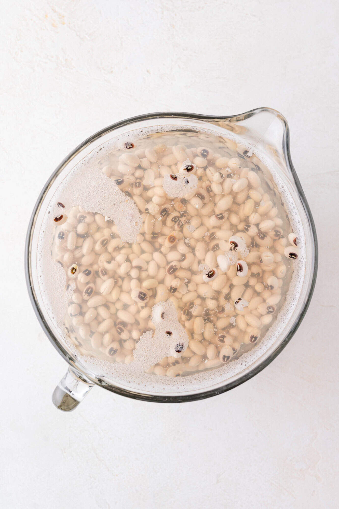 Black-Eyed Pea Soup 8 A glass measuring cup filled with water and soaking black-eyed peas, seen from above on a white surface.