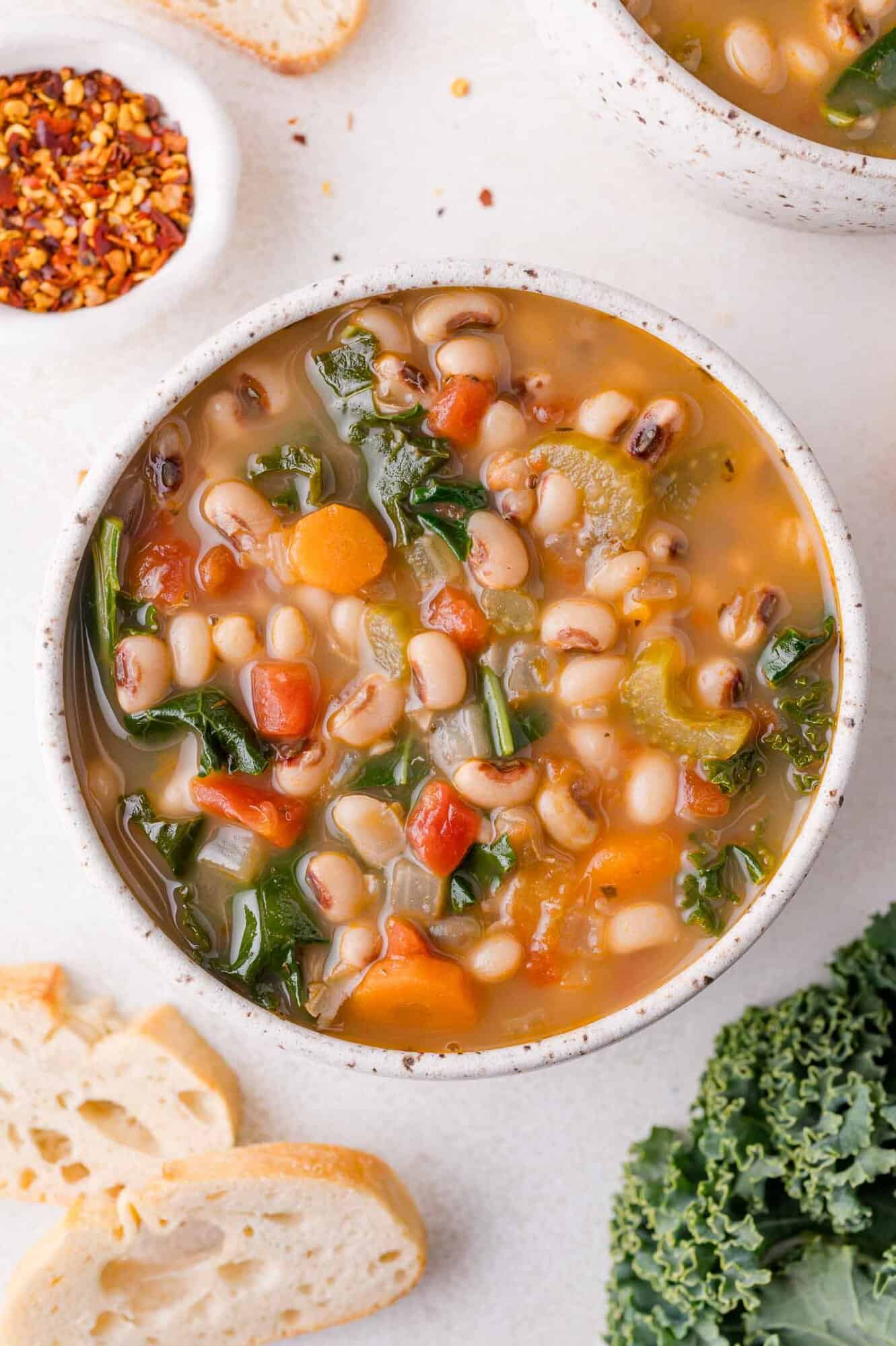 Black-Eyed Pea Soup 18 A bowl of black-eyed pea soup with tomatoes, greens, and carrots, accompanied by slices of bread and a small dish of red pepper flakes.
