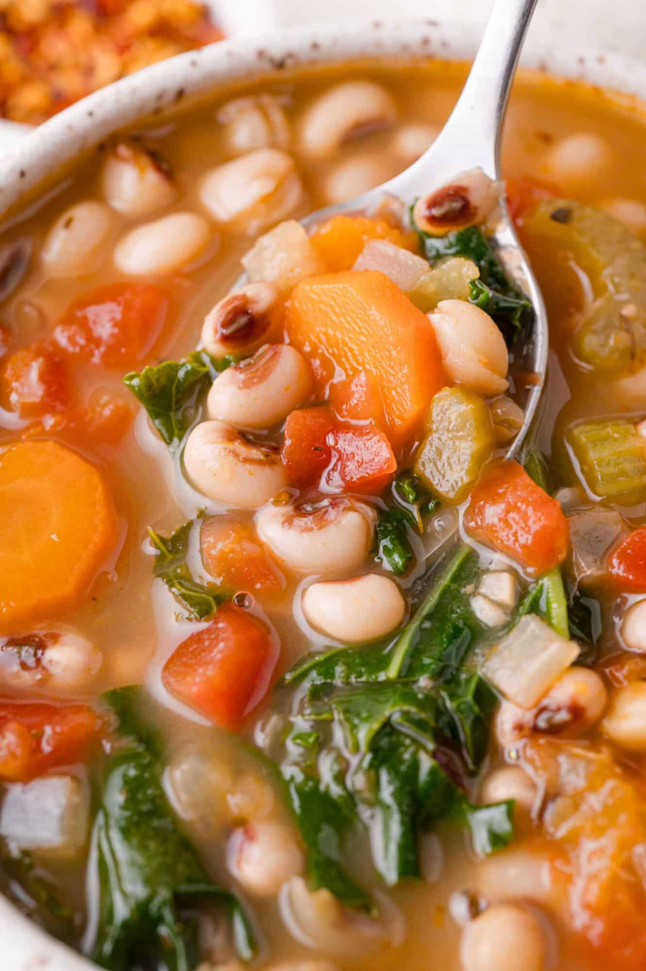 Black-Eyed Pea Soup 4 A close-up of a bowl of black-eyed pea soup with carrots, tomatoes, celery, and leafy greens, with a spoon lifting a portion.