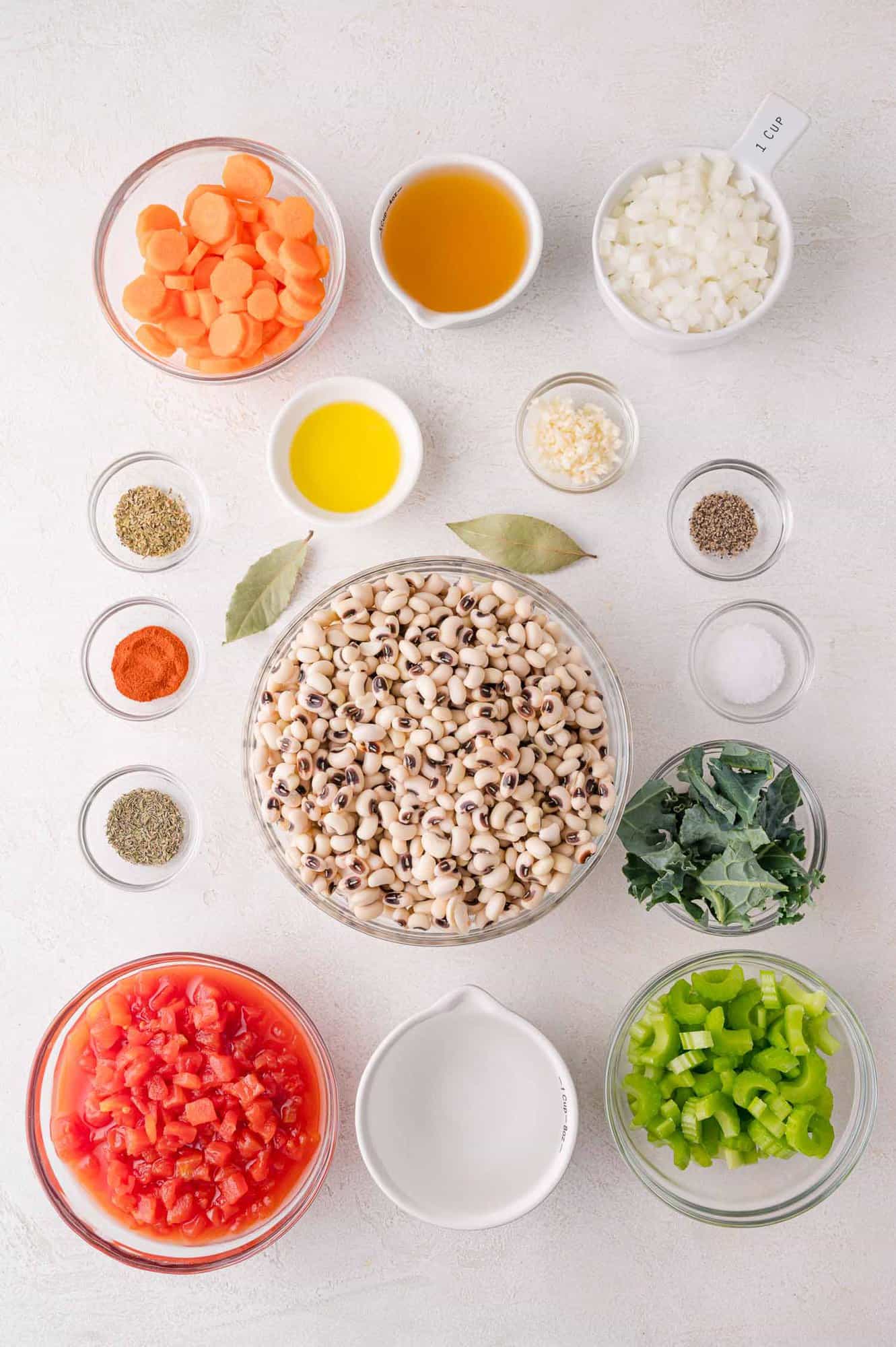 Black-Eyed Pea Soup 6 Overhead view of prepped ingredients in bowls, including black-eyed peas, diced tomatoes, carrots, celery, onion, garlic, kale, spices, bay leaves, broth, and oil on a white surface.