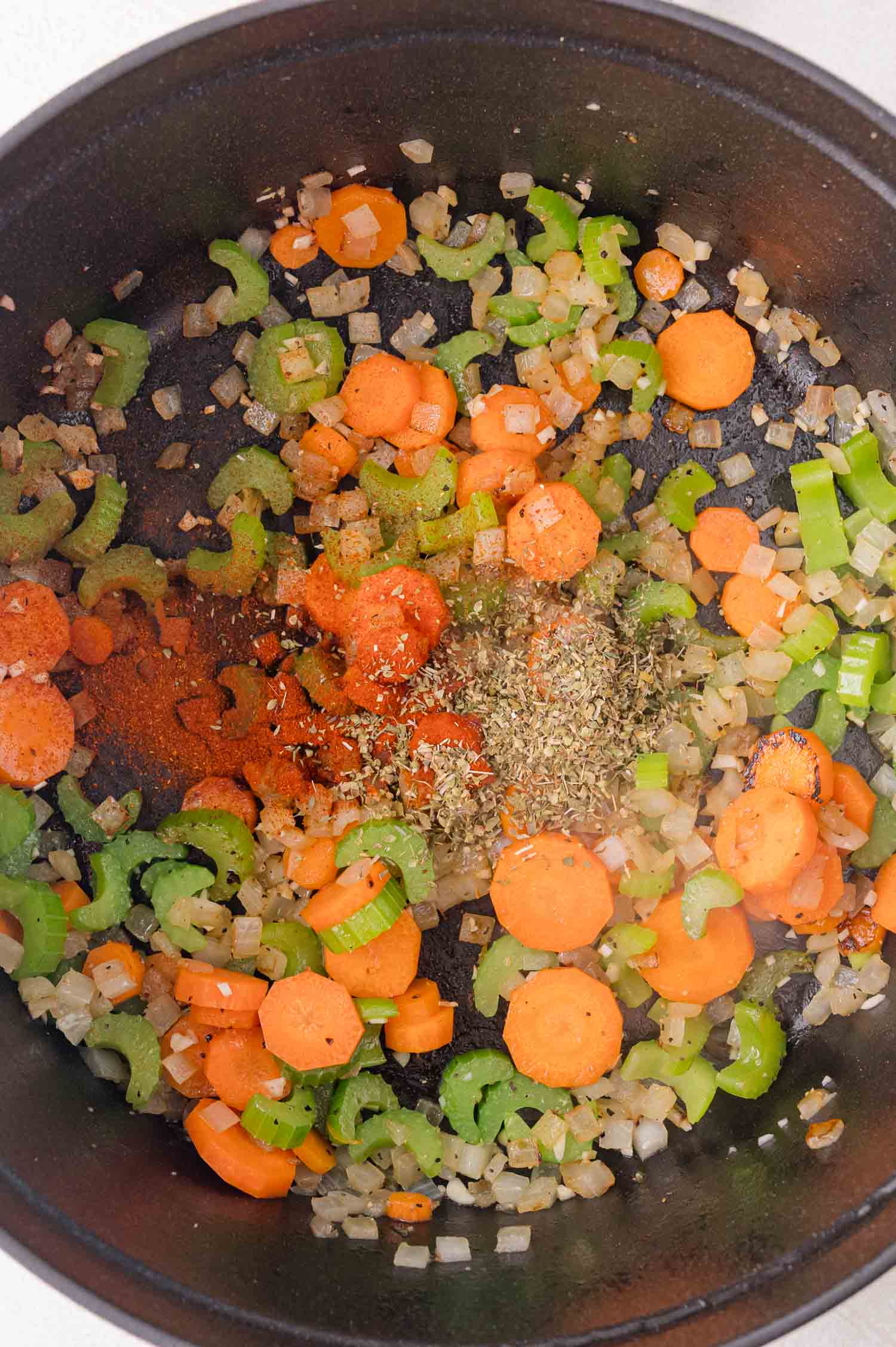 Chopped carrots, celery, and onions sautéing in a black pot with dried herbs and spices sprinkled on top.