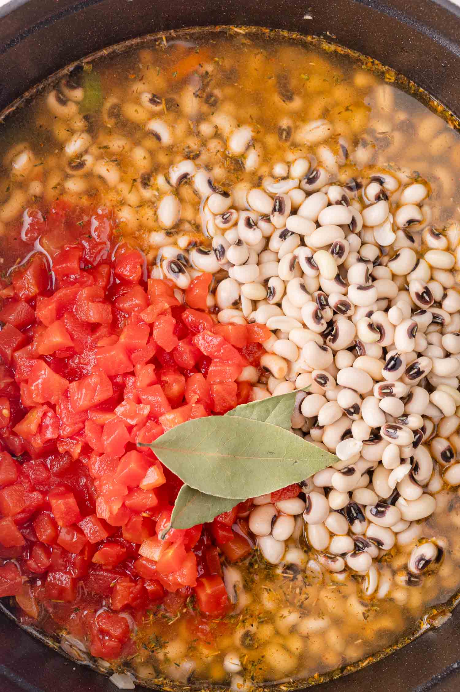 A pot containing black-eyed peas, diced tomatoes, seasonings, and two bay leaves in a simmering broth.