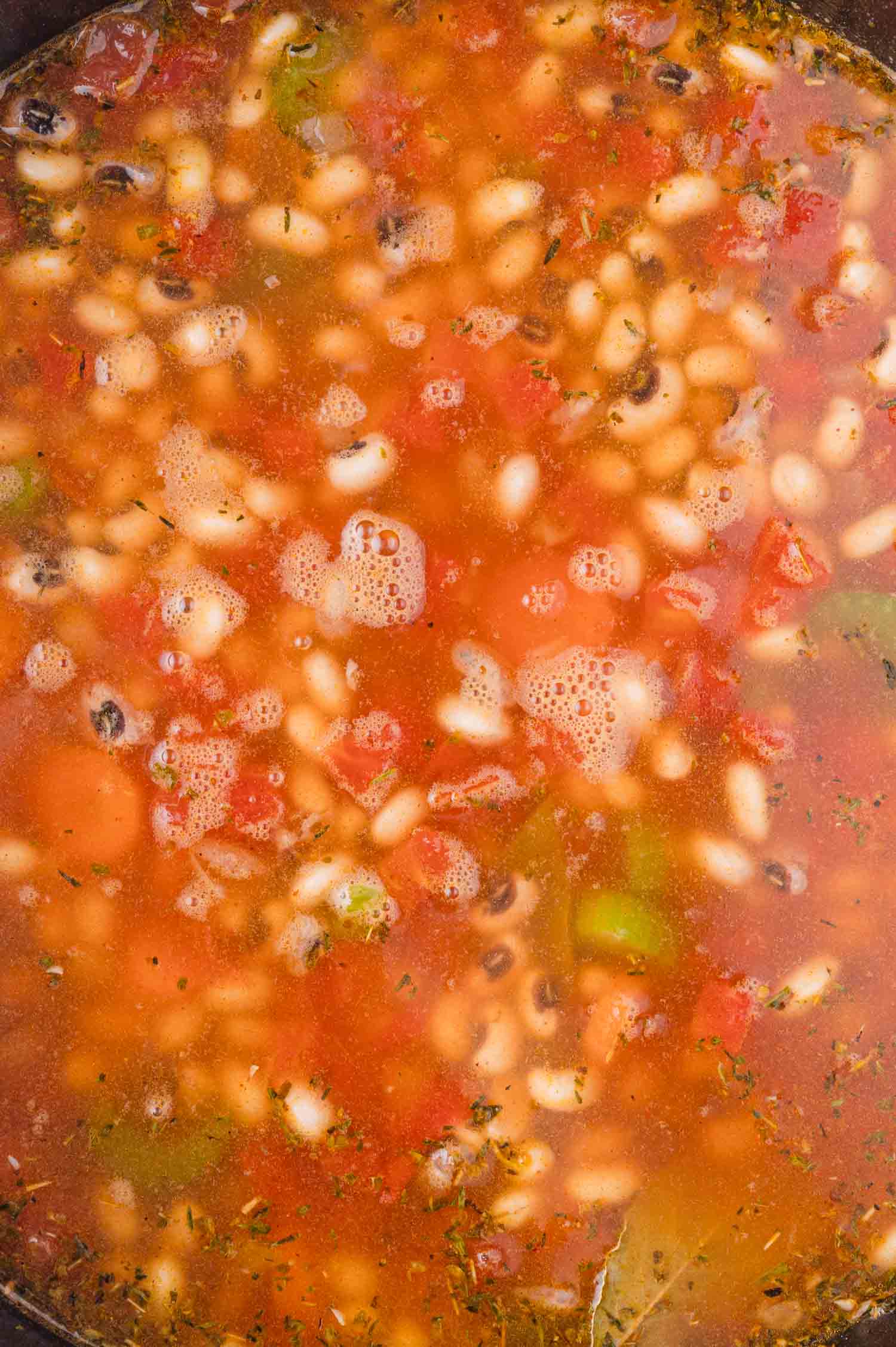 A close-up of a pot of stew with black-eyed peas, diced tomatoes, celery, and herbs in a red broth.