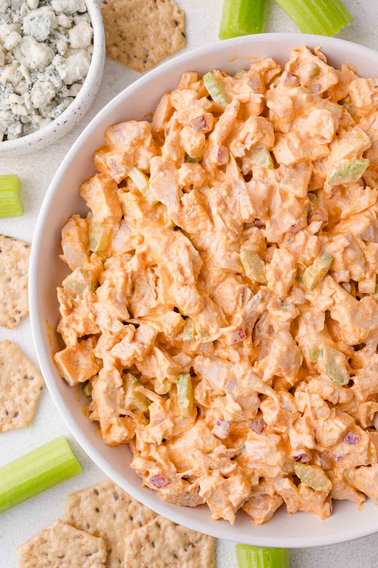 A bowl of buffalo chicken salad with diced chicken, celery, and creamy buffalo sauce, surrounded by celery sticks, crackers, and a bowl of blue cheese crumbles.