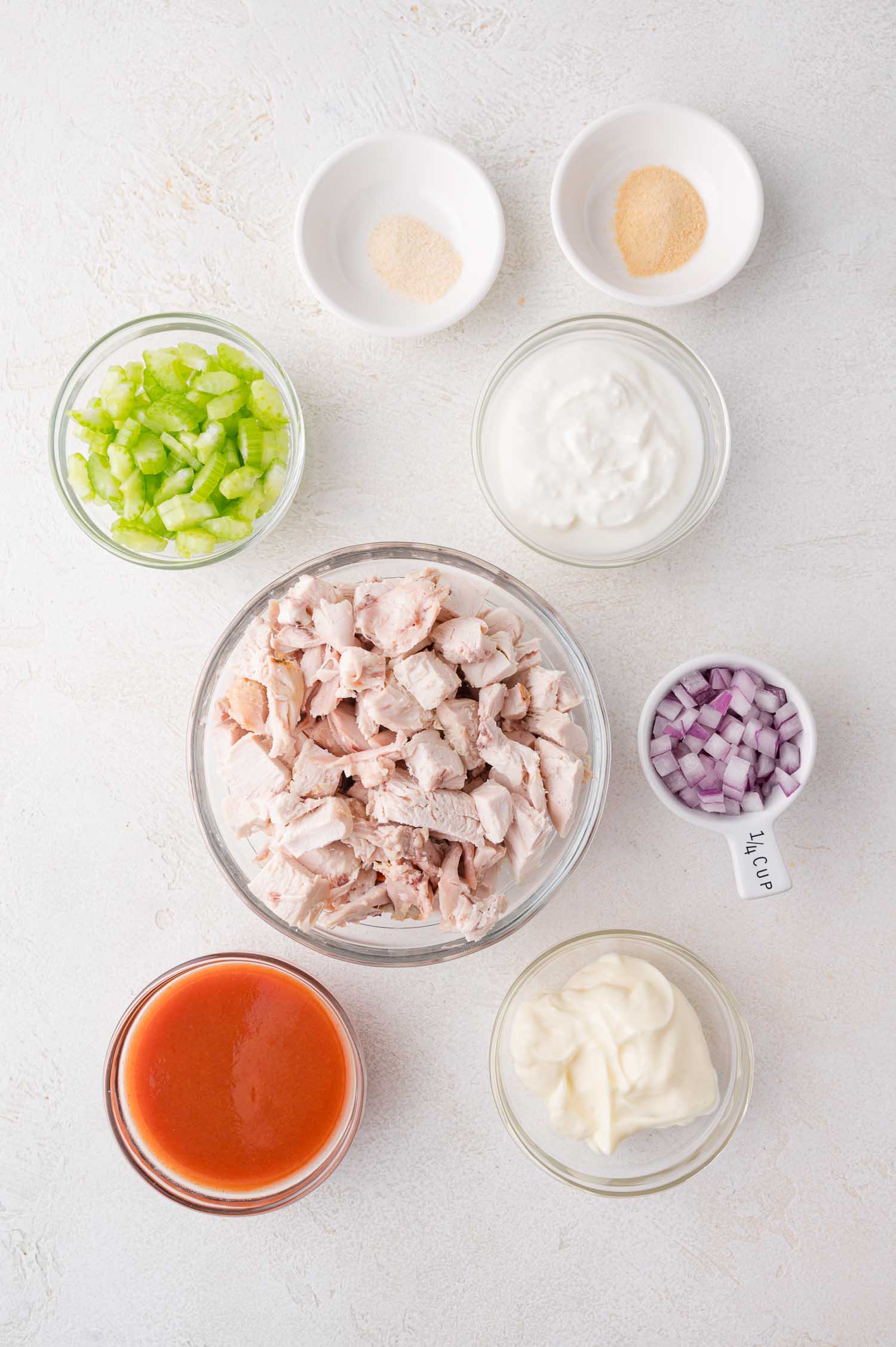 Seven bowls containing chopped celery, shredded cooked chicken, diced red onion, mayonnaise, yogurt, hot sauce, garlic powder, and onion powder arranged on a white surface.