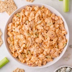 A bowl of buffalo chicken salad with chopped celery and crackers arranged around it on a light surface.