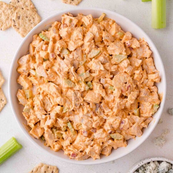 A bowl of buffalo chicken salad with chopped celery and crackers arranged around it on a light surface.