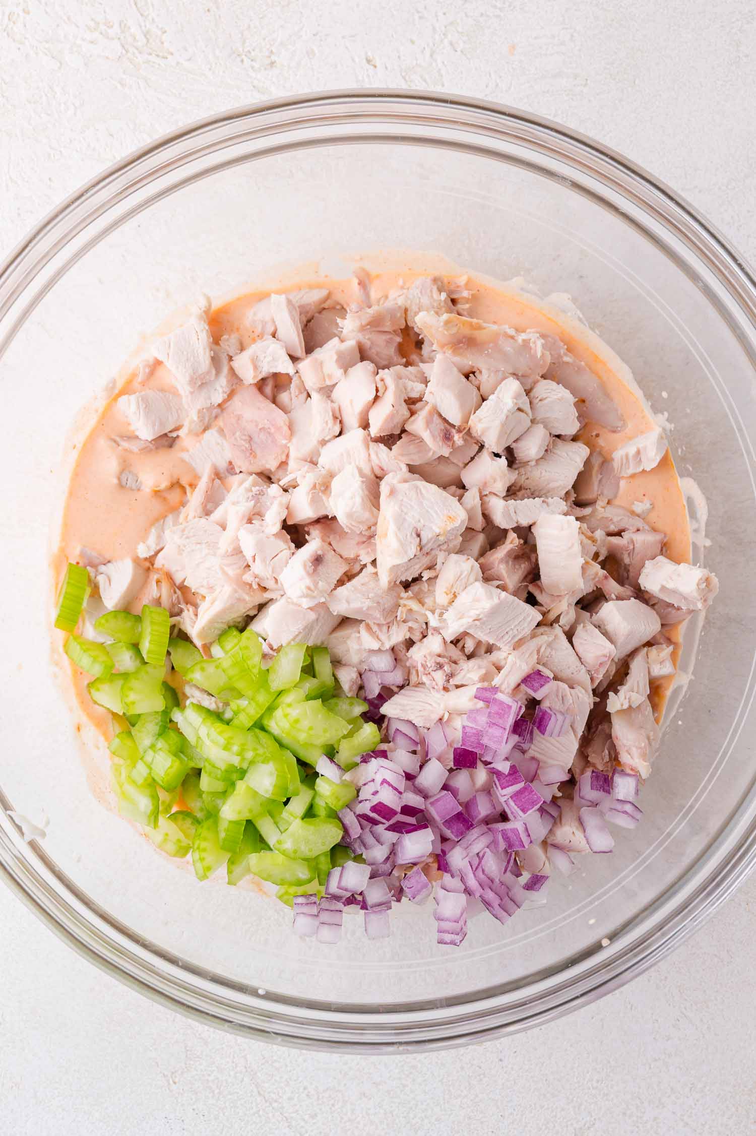A glass bowl containing chopped cooked chicken, sliced celery, and diced red onion over a creamy sauce.