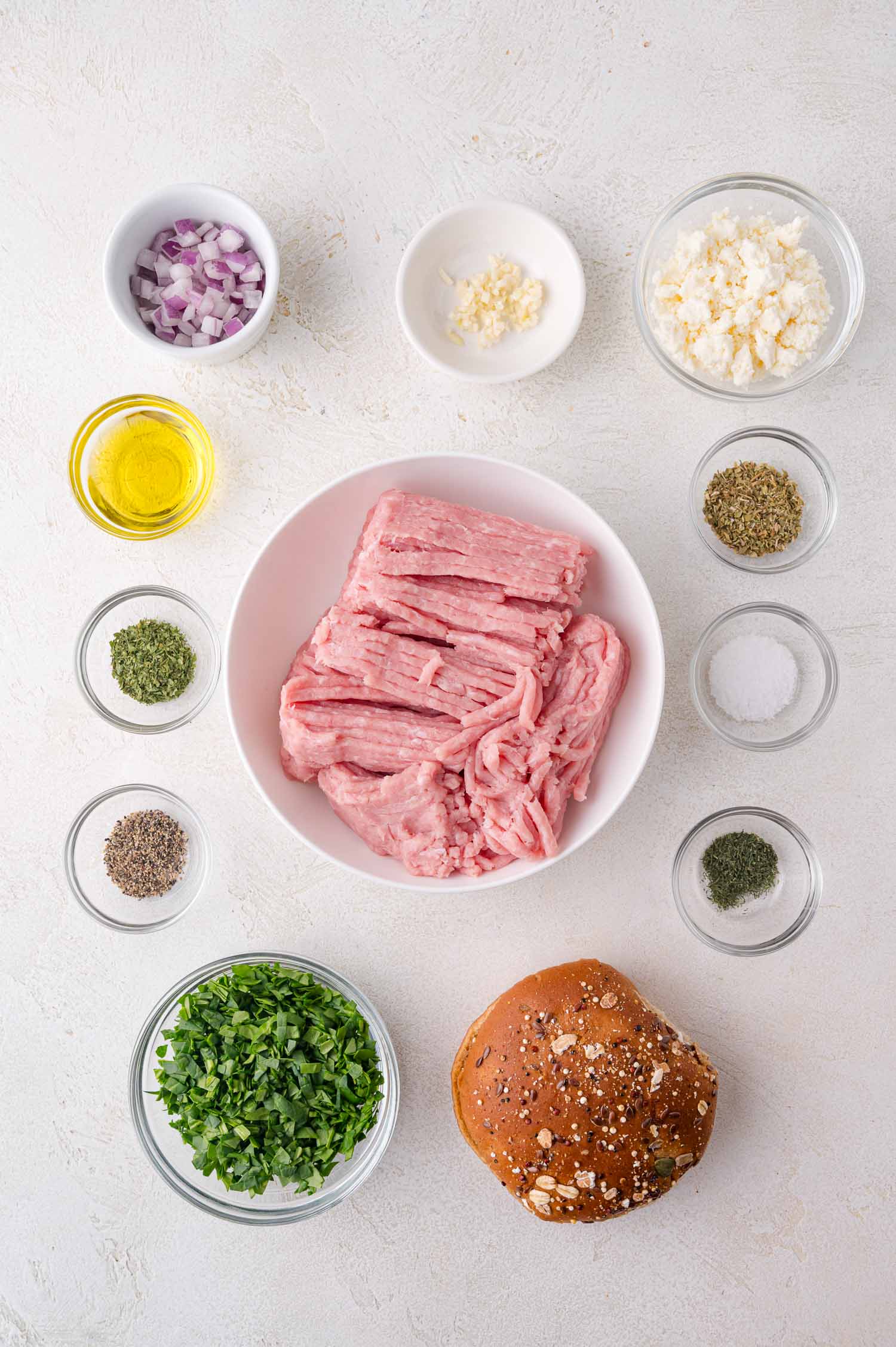 Ground turkey in a bowl surrounded by small bowls of chopped onion, garlic, parsley, feta, spices, olive oil, and an oat-topped burger bun on a white surface.