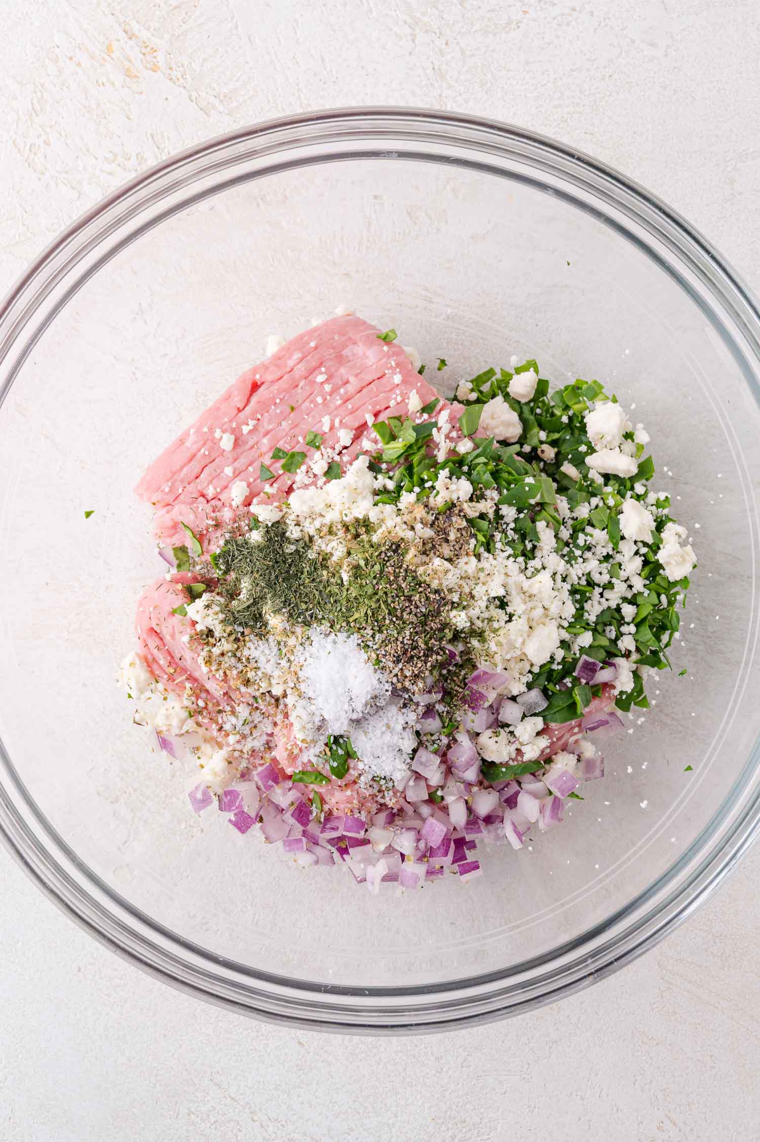 Ground meat, chopped red onions, herbs, spices, salt, and crumbled cheese are arranged in a clear glass mixing bowl on a light surface.