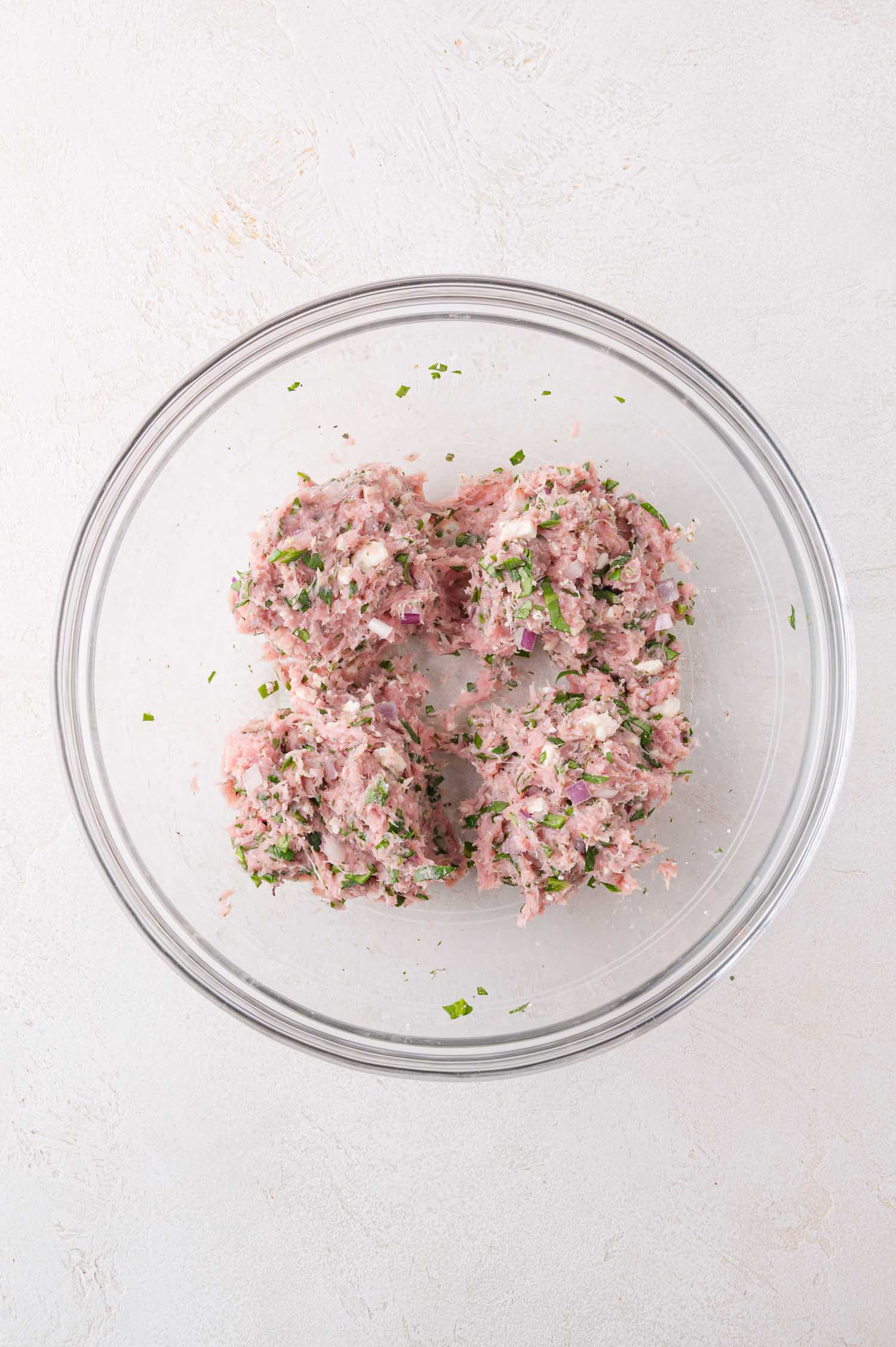 A glass bowl containing a mixture of ground meat, herbs, and chopped onions divided into four portions on a light surface.