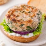 A turkey burger patty with herbs on a bun, layered with green leaf lettuce, sliced red onion, and white sauce, served open-faced on a plate.