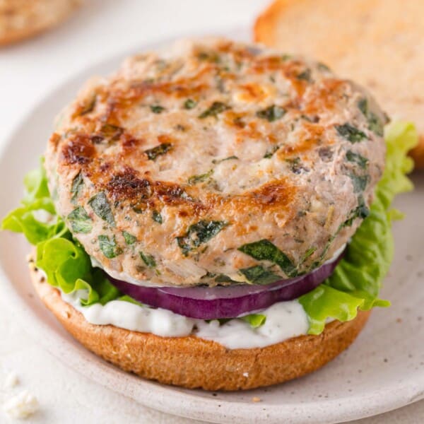 A turkey burger patty with herbs on a bun, layered with green leaf lettuce, sliced red onion, and white sauce, served open-faced on a plate.