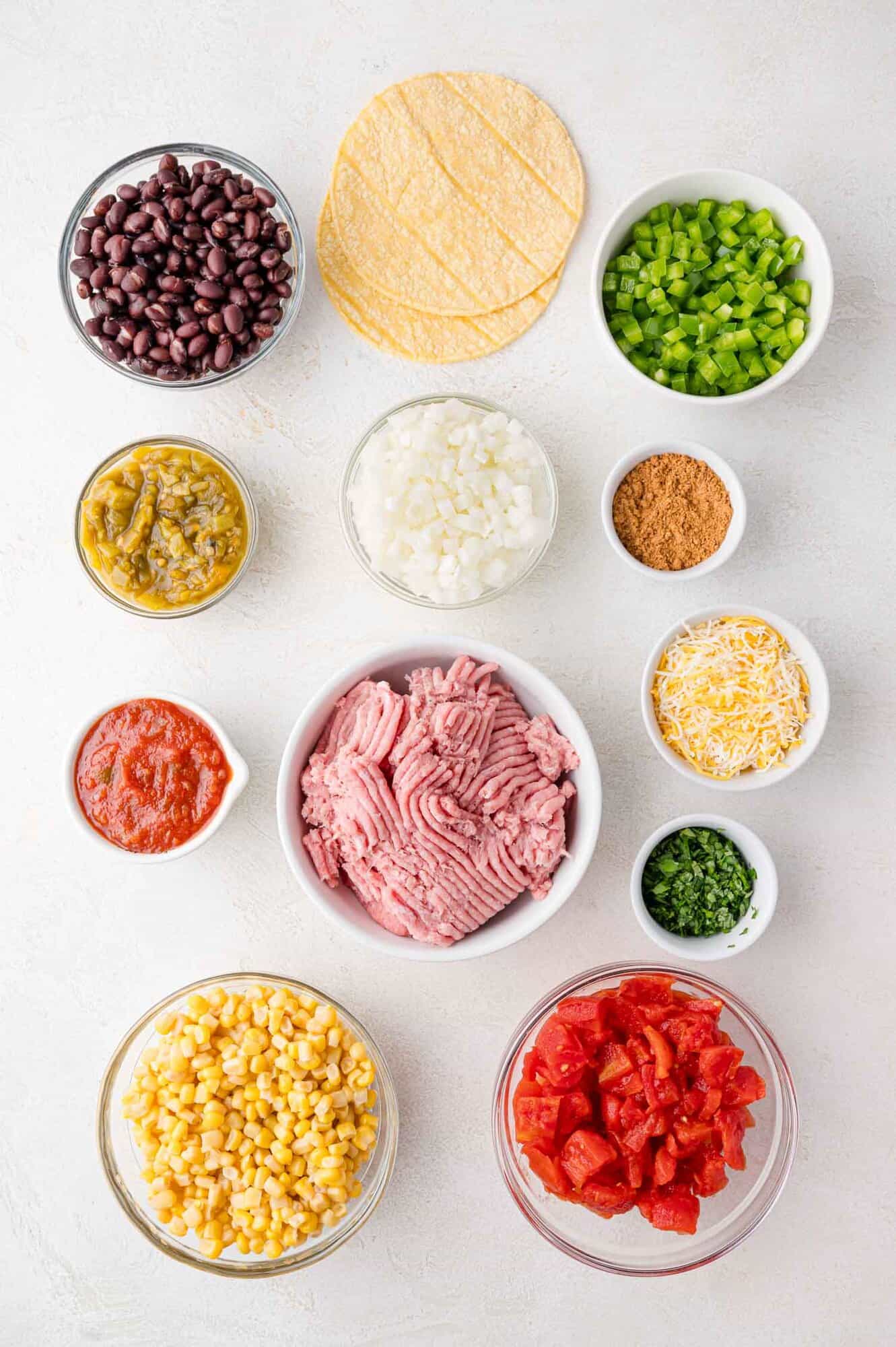 Assorted ingredients, including ground meat, black beans, corn, diced tomatoes, onions, green chilies, cheese, salsa, bell pepper, seasoning, herbs, and corn tortillas, arranged on a white surface.