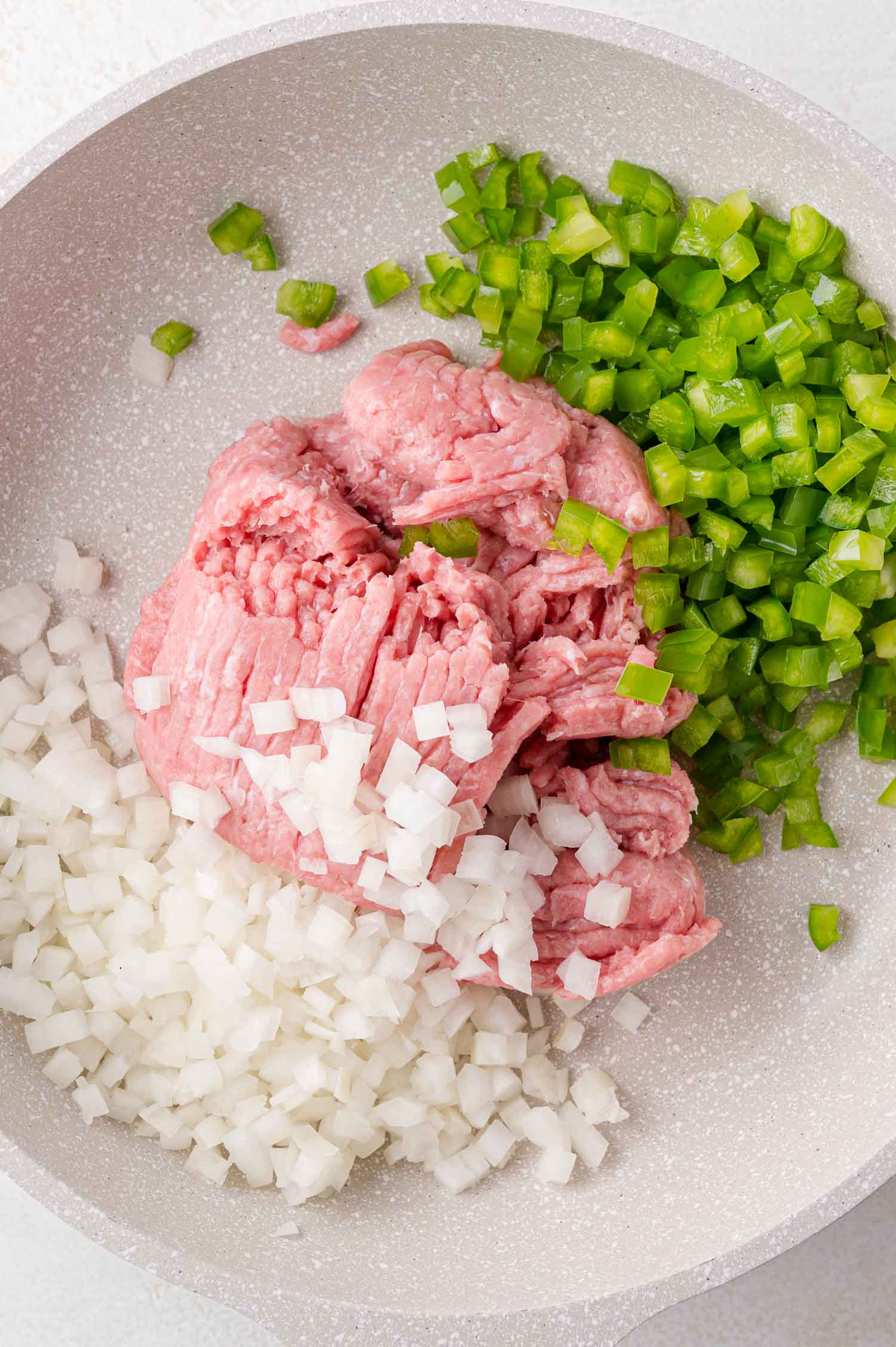 Raw ground meat, chopped white onions, and diced green bell peppers in a white frying pan, ready to be cooked.